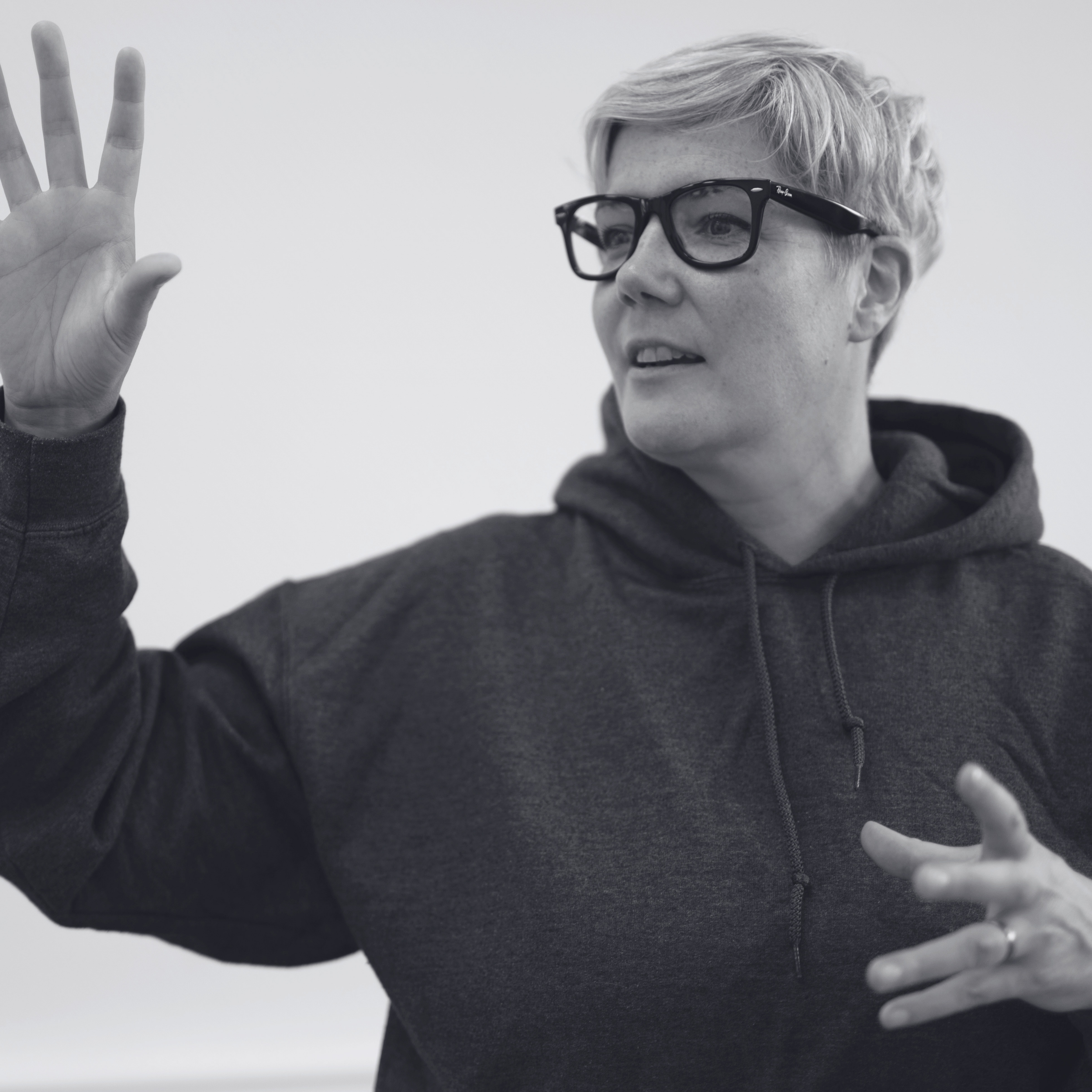 A woman with short, wavy silver hair, wearing large black glasses and a dark hoodie, raising her right hand with an open palm.