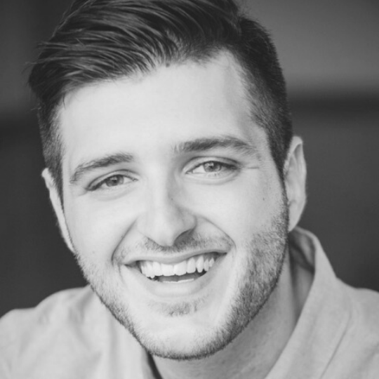 [Image Description: Black and white headshot of a man with short, slicked back hair and short facial hair. Frank smiles widely at the camera.]