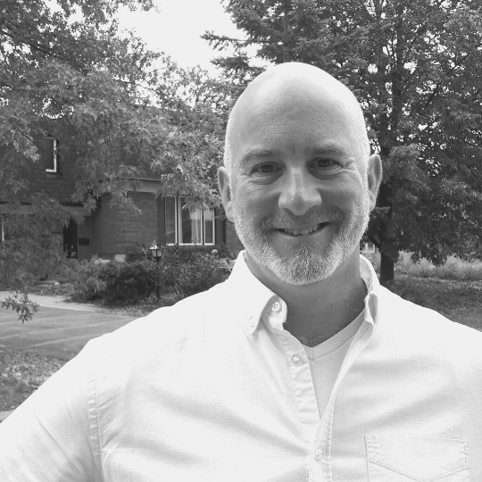 [Image description: Black and white headshot of a man wearing a button up shirt. Sean has a shaved head, a short beard, and is smiling, standing in front of trees and a house.]