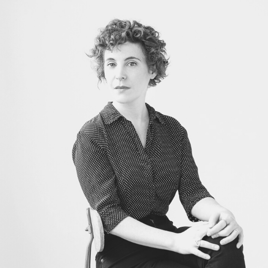 [Image Description: Black and white headshot of a short curly haired person, Sarah, wearing a collared shirt with the top two buttons undone. They are sitting with one leg crossed over the other, hands interlaced on top.]