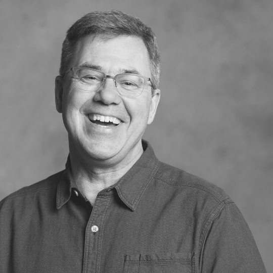 [Image Description: A black and white headshot of a short haired man with frameless glasses. He has a button up shirt with the top button undone. Eric smiles widely with his teeth showing.]