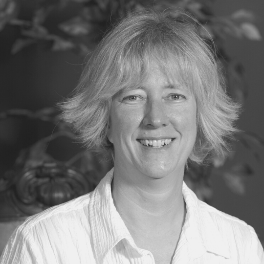 [Image Description: Black and white headshot of a woman with short hair and bangs. Jane is wearing a button up shirt with a collar. She is smiling widely at the camera.]