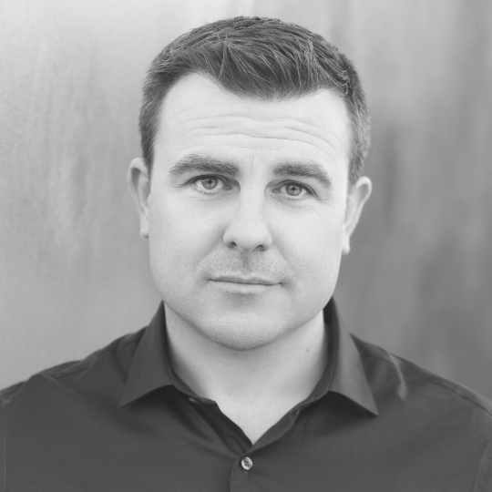 [Image Description: Black and white headshot of a man with short hair. Brad is wearing a button up shirt with a collar and is looking at the camera.]