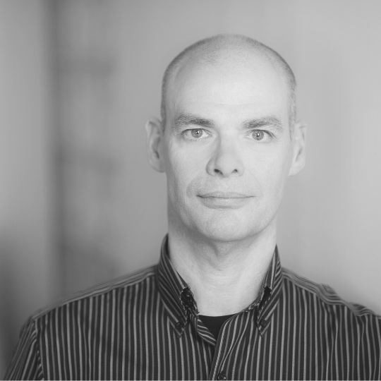 [Image Description: Black and white photo of a man looking at the camera. Geoff has very short shaved hair and is wearing a striped button up shirt with a collar.]