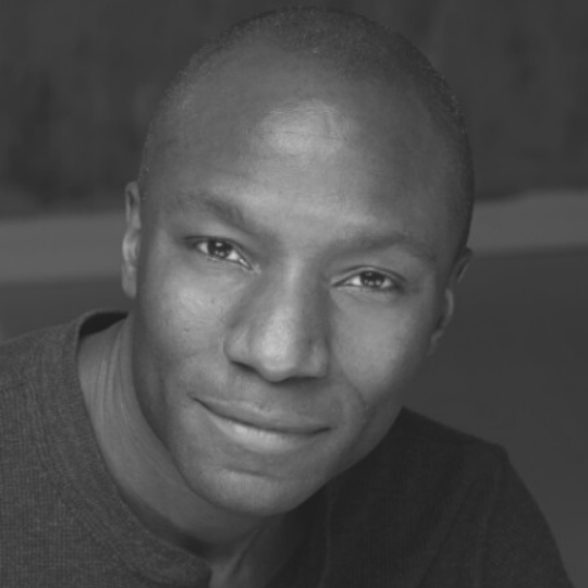 [Image Description: Black and white headshot of a man smiling softly at the camera. Andrew has a shaved head, is wearing a plain shirt, and is leaning forward.]