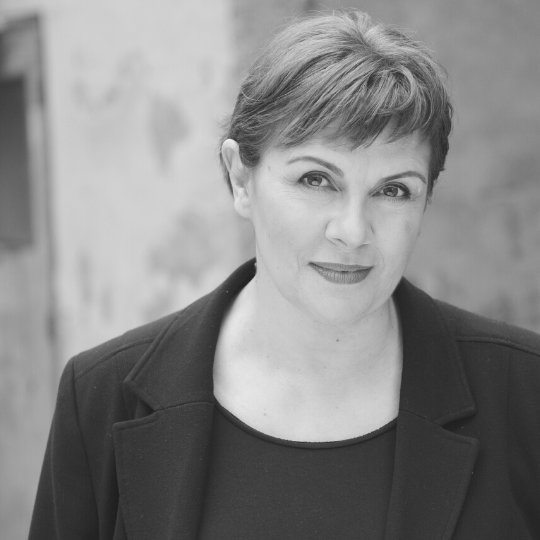 [Image Description: Black and white headshot of a woman with short cropped hair and bangs. Mary is wearing a blazer and is smiling softly at the camera in front of a wall with a door to the right.]