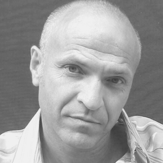 [Image Description: Black and white photo of a man with very short hair, wearing a collared shirt. Roger’s head is tilted to the left as he looks at the camera.]