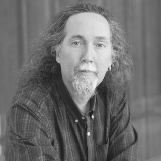 [Image Descriptions: Black and white headshot of a man with long, curly hair with a mustache and a goatee. Peter is wearing a plaid button-up shirt and is leaning towards the camera.]