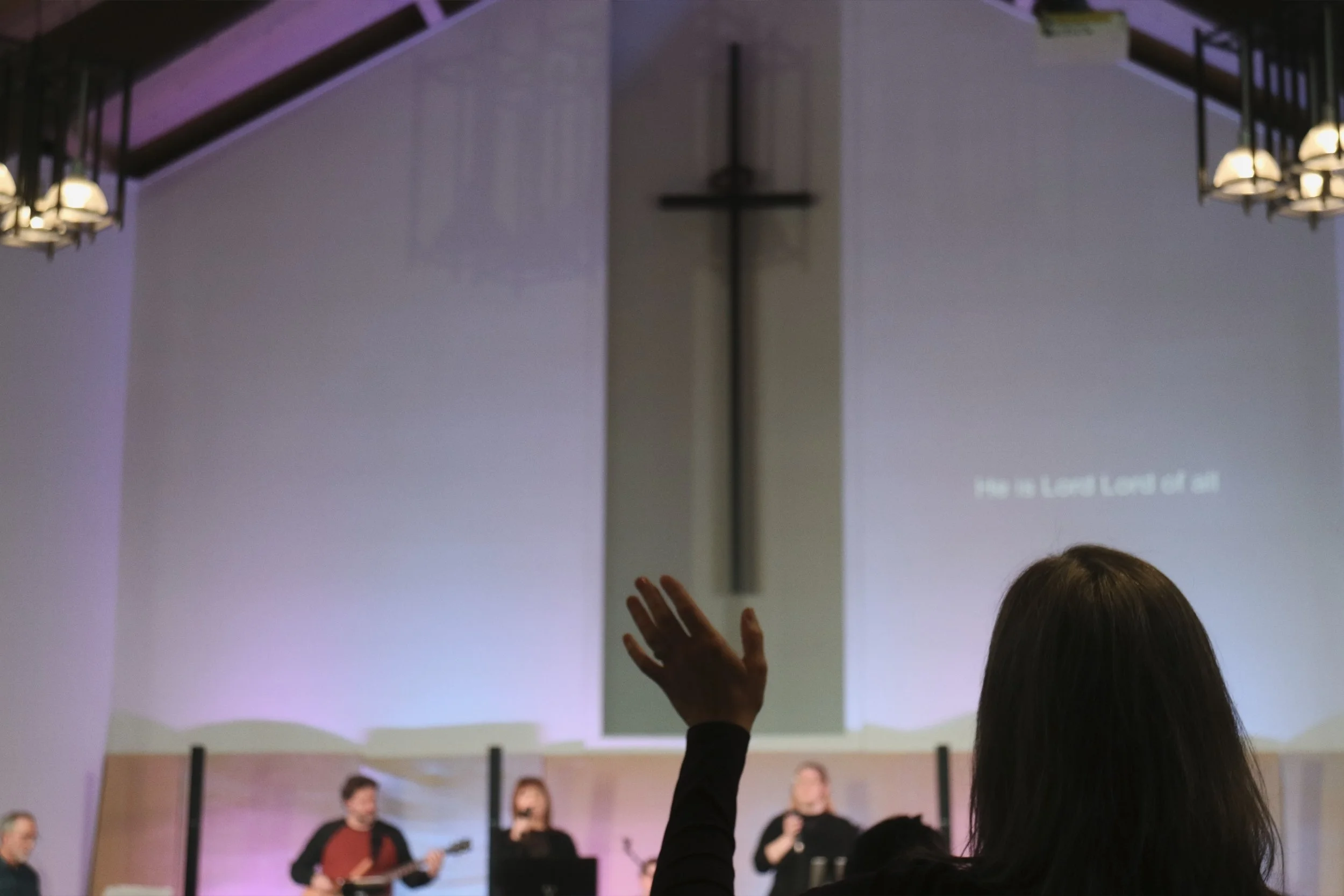 A woman raising her hand during a church service, with a cross on the wall and a projected song lyric in the background, while several people sing and play instruments.