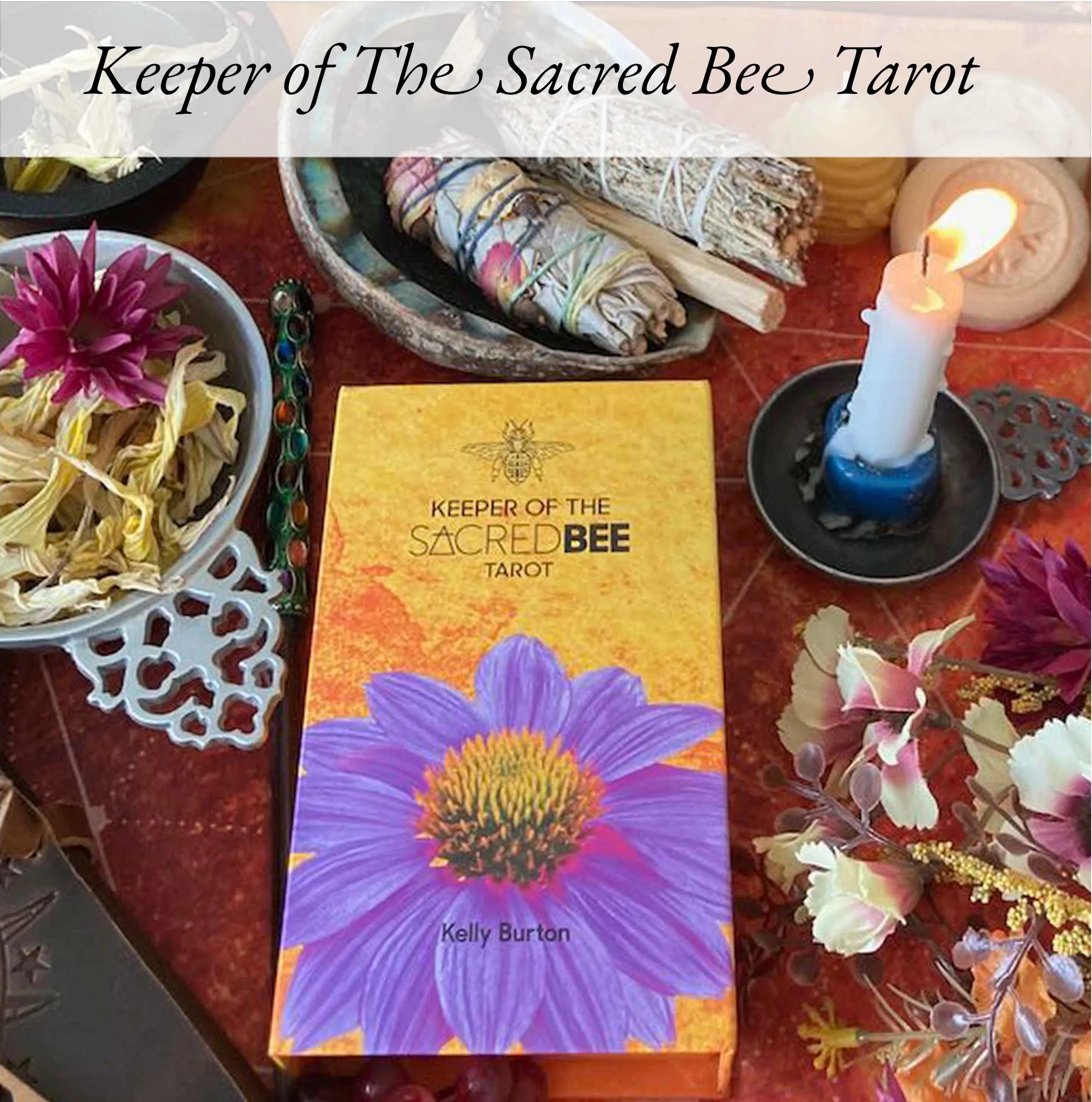 The image shows the book titled 'Keeper of the Sacred Bee Tarot' by Kelly Burton, placed on a table surrounded by flowers, candles, and tarot reading materials. The cover features a large purple flower and a bee illustration.