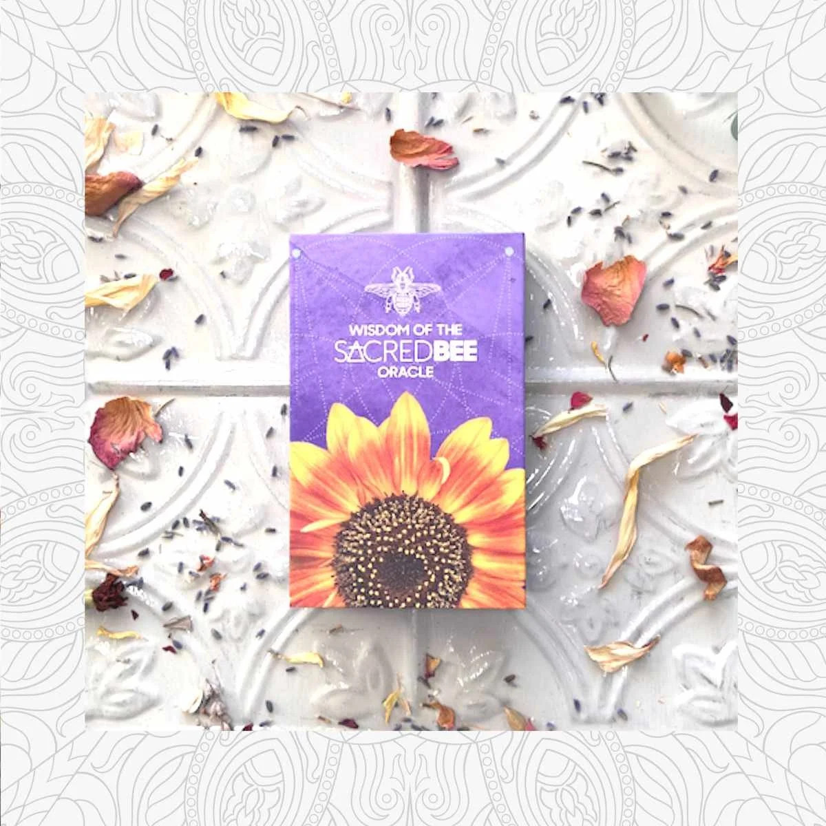 A purple card with a sunflower and bee illustration, titled 'Wisdom of the Sacred Bee Oracle,' placed on a decorative white surface surrounded by dried flower petals and small dried herbs.