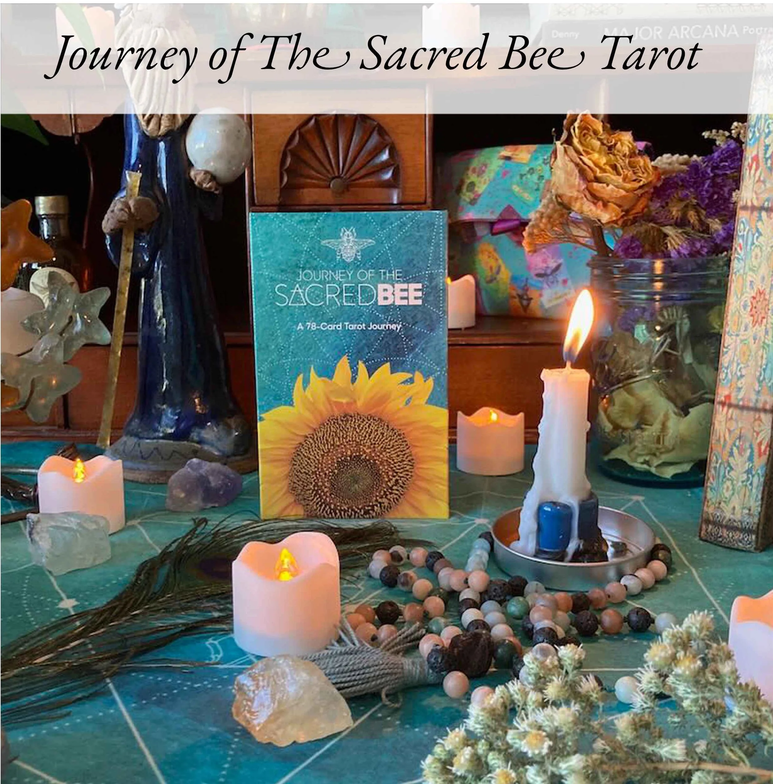 An arrangement of tarot and spiritual items on a tabletop, including a card deck titled 'Journey of The Sacred Bee Tarot,' candles, crystals, dried flowers, and a string of beads, set against a background with a colorful cloth and decorative objects.