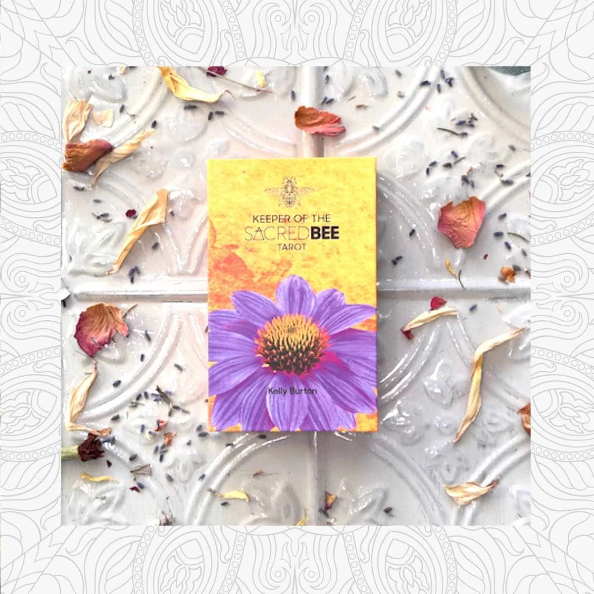 Book titled 'Keeper of the Sacred Bee Tarot' by Kelly Burton, placed on a decorative surface with dried flower petals and small black seeds scattered around it.