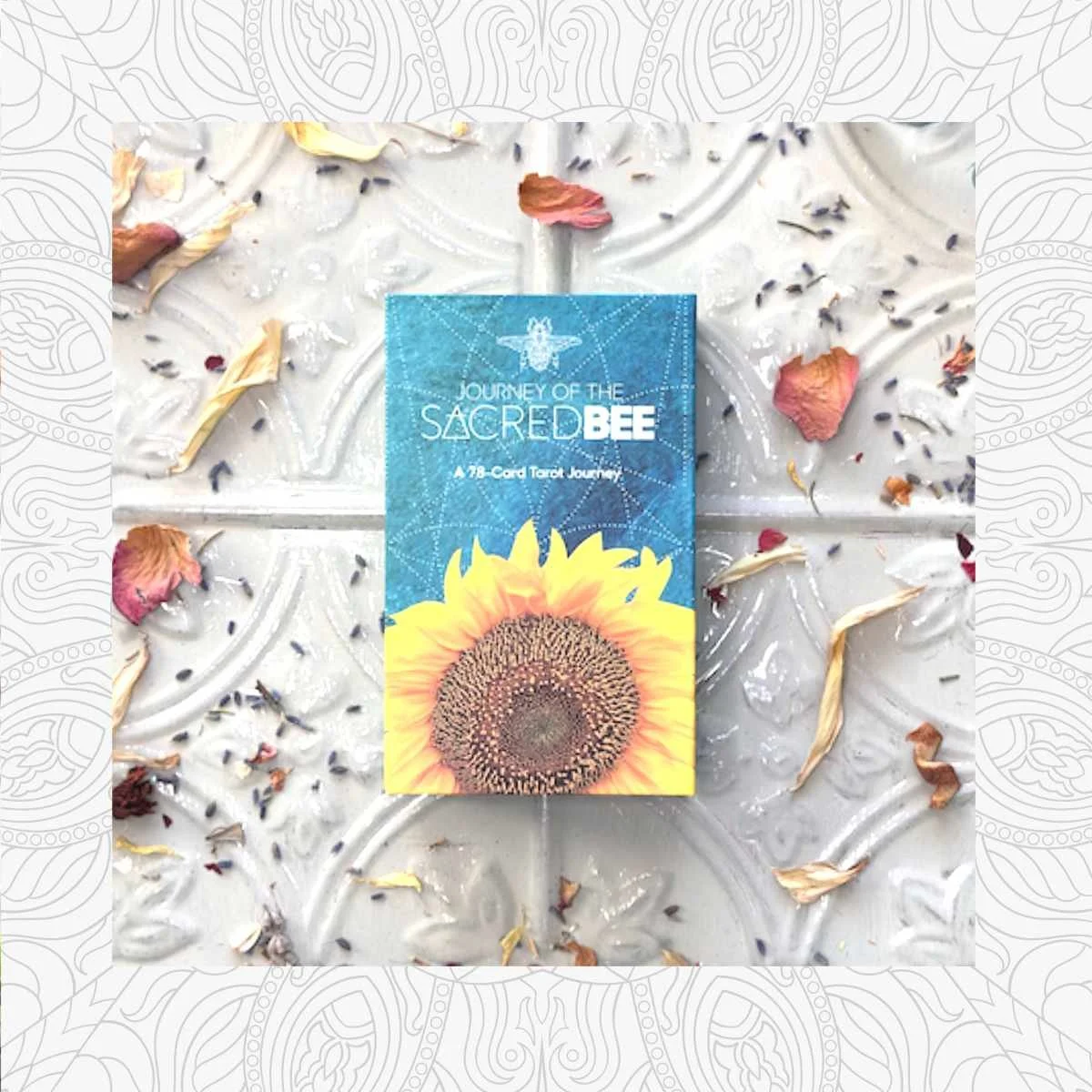 A card titled "Journey of the Sacred Bee" with a sunflower on the cover, surrounded by dried flower petals and lavender on a decorative white surface.