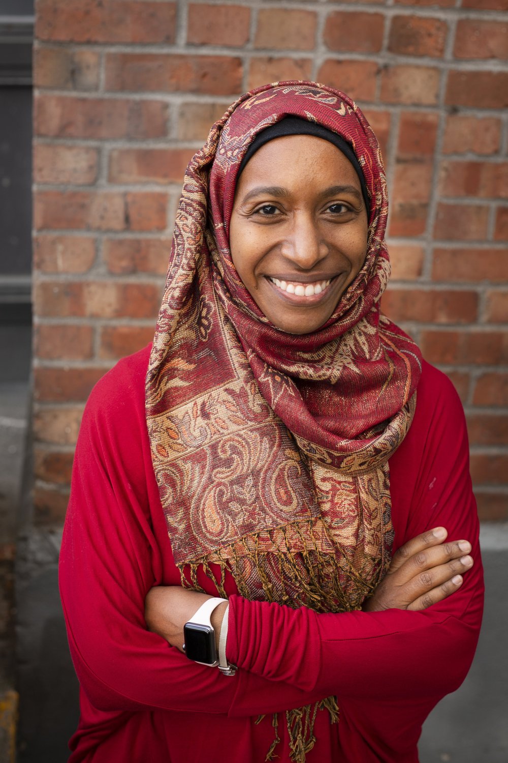 Photograph of Nasreen Al-Tamimi smiling with her arms crossed. She wears a red headscarf that matches her shirt.