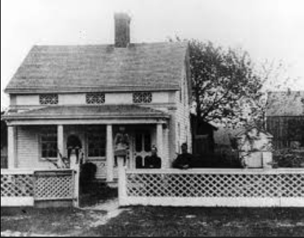 Black and white photo of a two-story house with a porch, decorative railing, and chimney on the roof, surrounded by trees and a small yard.