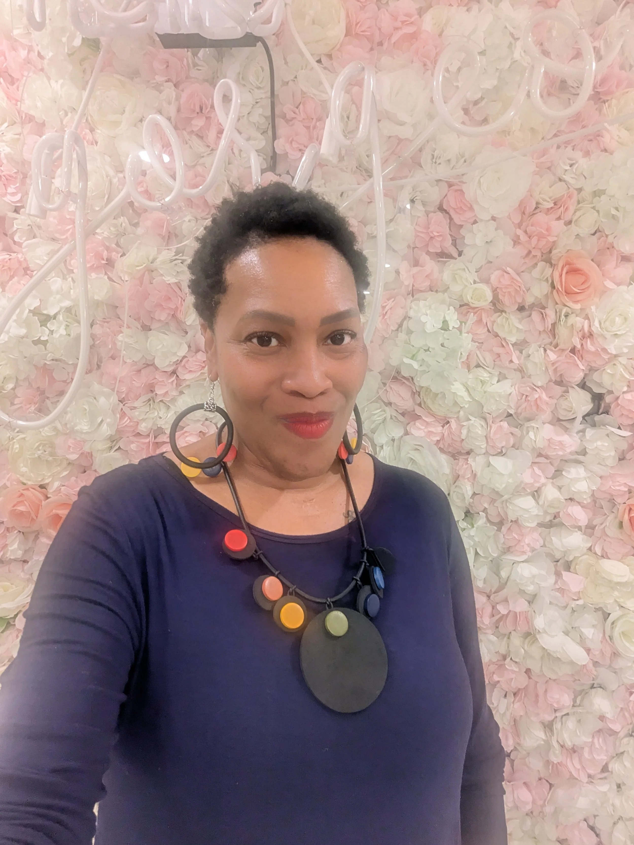 A woman with short curly black hair and red lipstick taking a selfie in front of a pink and white floral wall, wearing a navy blue top and a colorful beaded necklace.