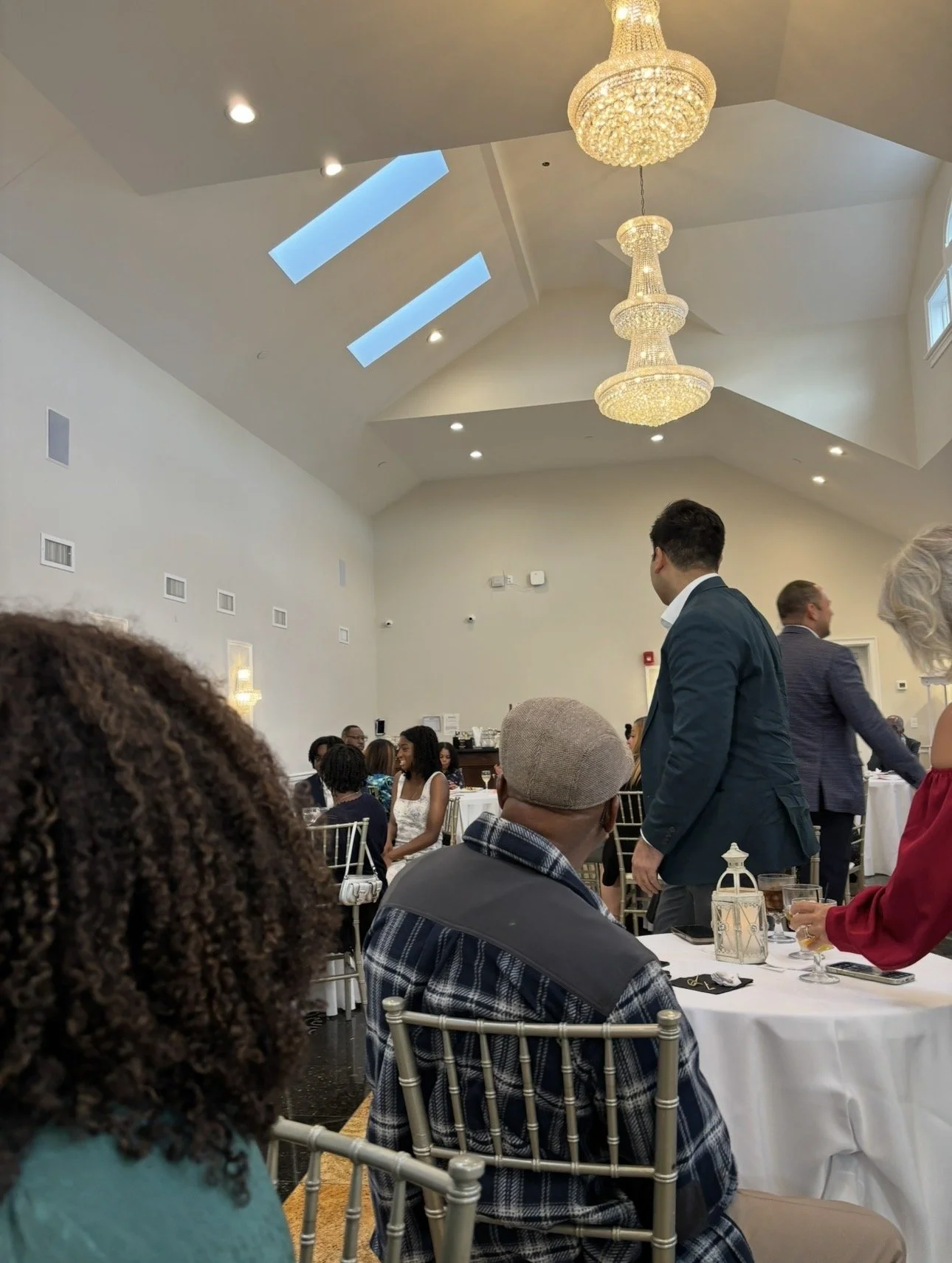 A banquet hall filled with guests seated at tables, with some standing and socializing. The hall has high ceilings with skylights, elegant chandeliers, white walls, and decorative lanterns on the tables. The people are dressed in formal attire, sugge