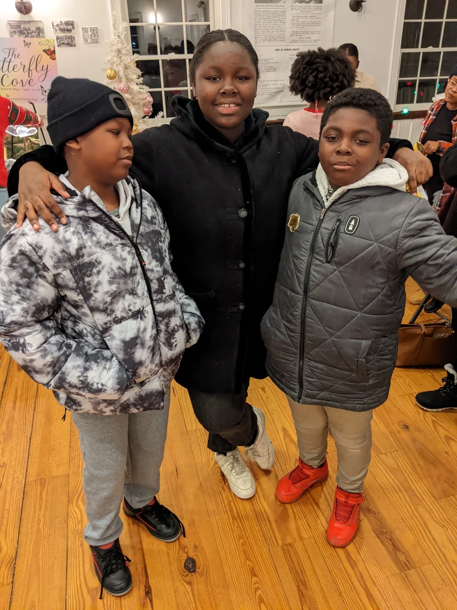 Three children standing with an adult woman at a holiday gathering. The girl on the left wears a black beanie and camouflage jacket. The girl in the middle wears a black coat. The boy on the right wears a gray winter jacket and bright red sneakers. C
