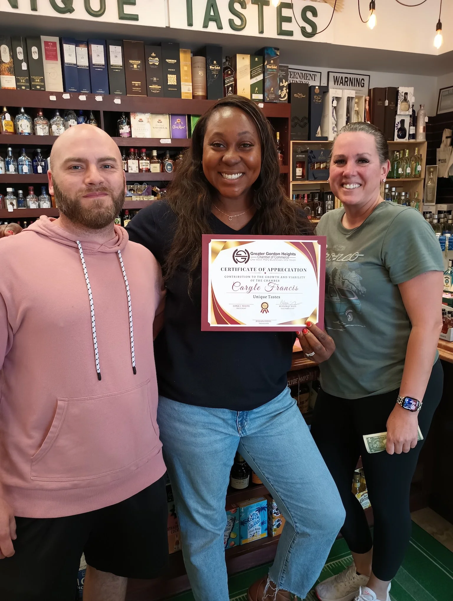 Three people standing inside a liquor store, smiling and holding a certificate of appreciation. Behind them are shelves filled with bottles of alcohol and liquor boxes, with a sign above reading ''Unique Tastes.'' The person in the middle is holding 