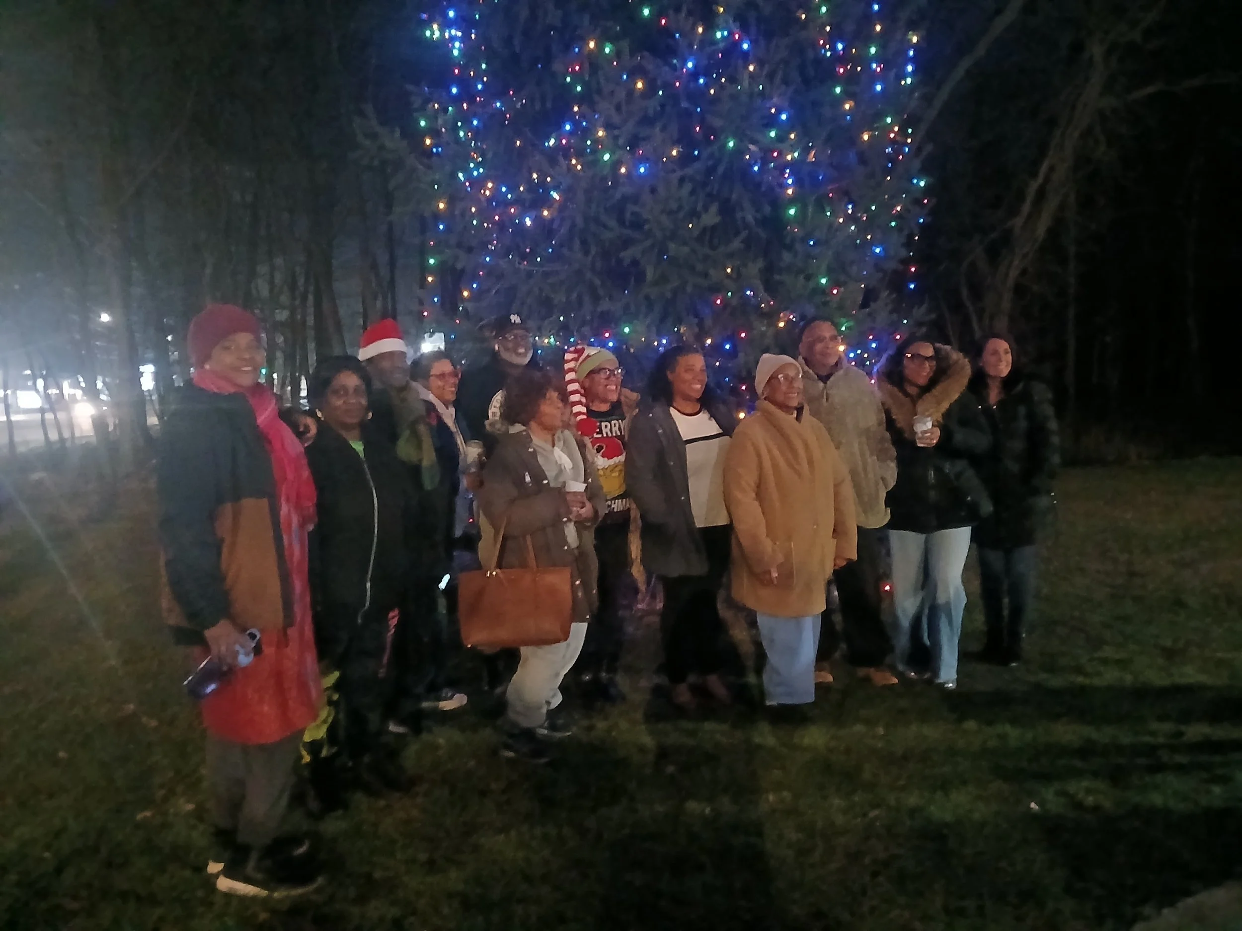 A group of people standing outdoors at night in front of a large Christmas tree decorated with multicolored lights. Several individuals are wearing Christmas-themed clothing, such as Santa hat, sweater, or scarf, and some are smiling or laughing.