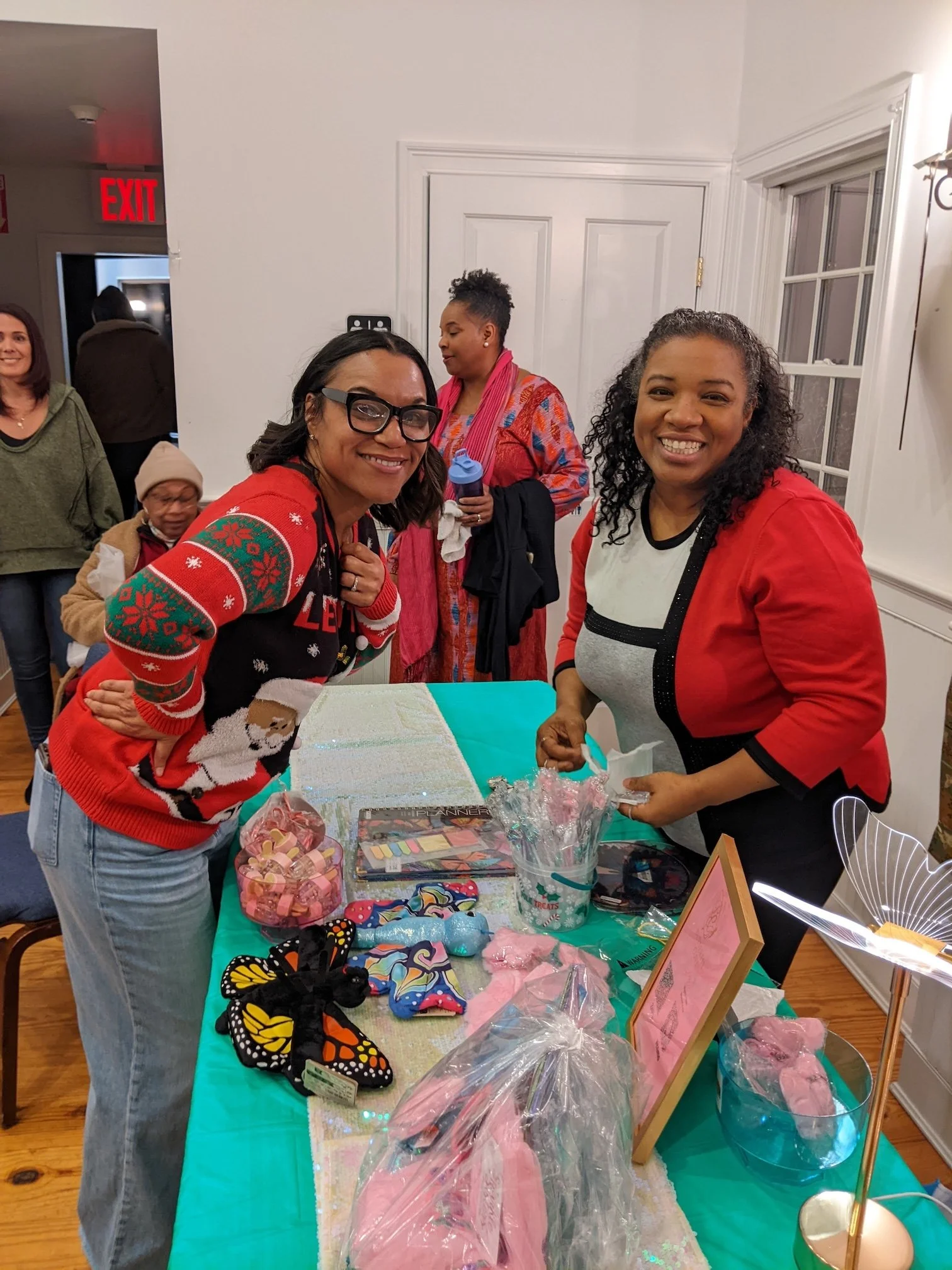 Group of women smiling at a craft or gift table with butterfly crafts, pink fabric, and pink marshmallow treats. One woman in front wears glasses and a Christmas sweater, another woman on the right wears a red jacket, and others are in the background