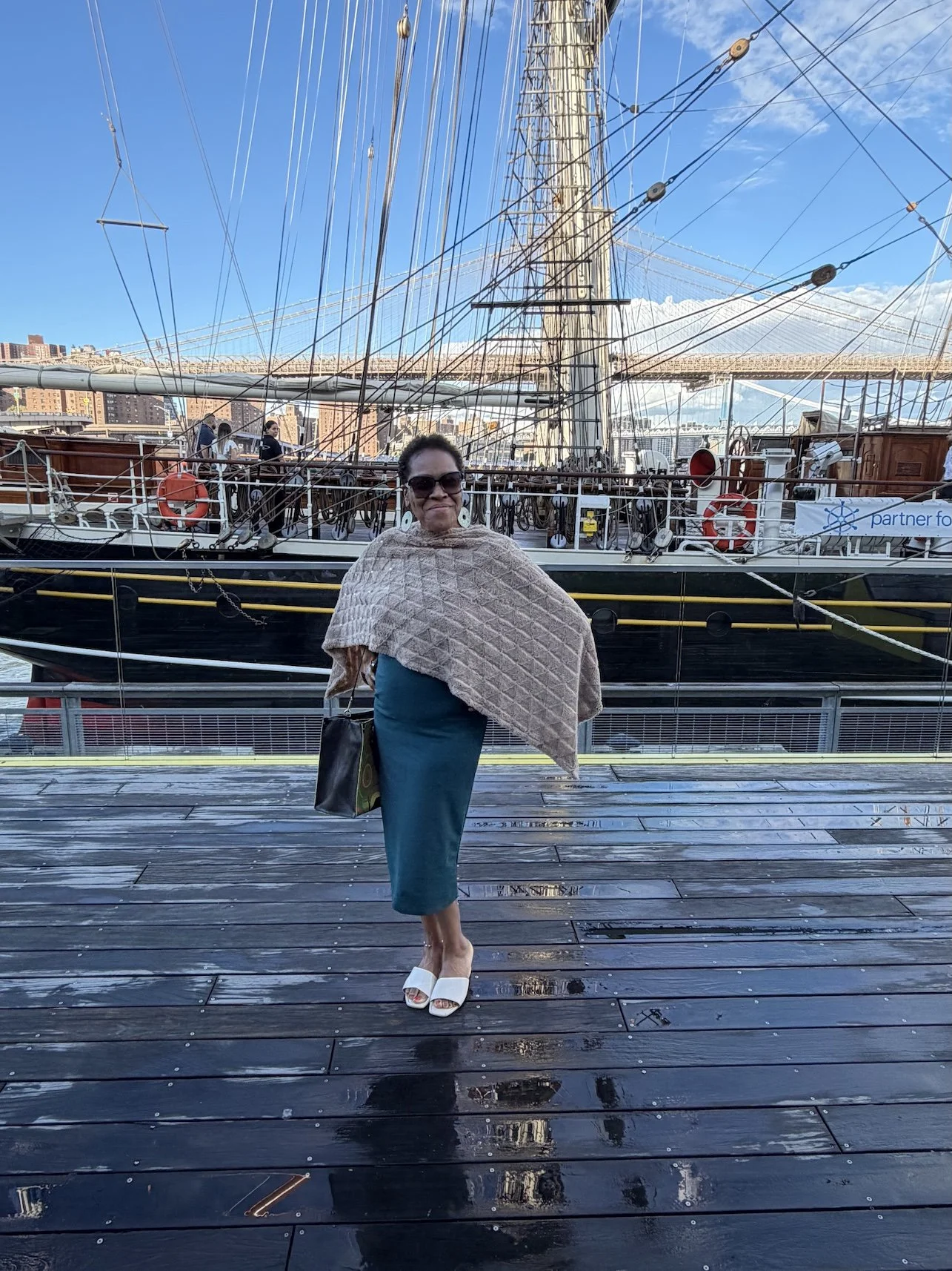 A woman wearing sunglasses, a teal dress, white shoes, and a beige shawl standing on a wet wooden dock in front of a tall ship with numerous ropes and riggings, with city buildings and a blue sky with some clouds in the background.