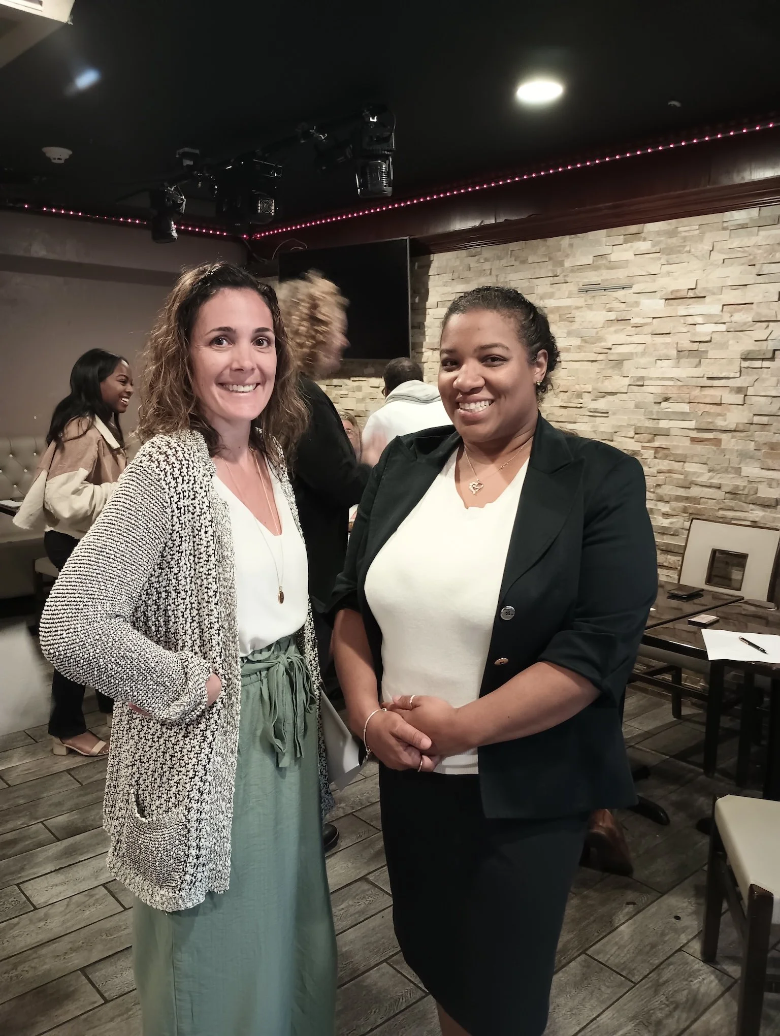 Two women standing close together, smiling at the camera, in a restaurant or cafe with a brick accent wall, surrounded by other people.