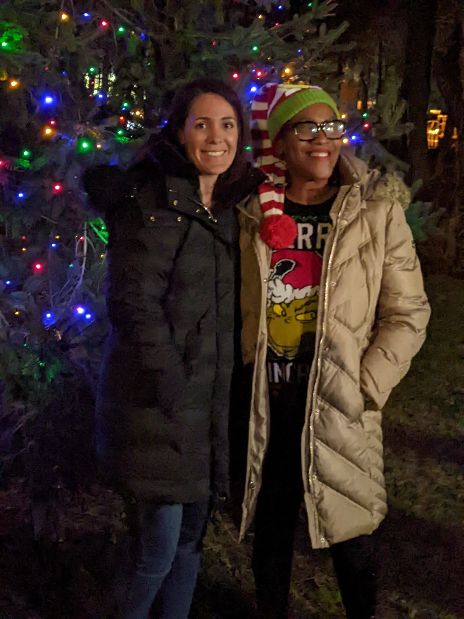 Two women standing in front of a Christmas tree with colorful lights. One is wearing a dark jacket and the other is wearing a tan coat and a festive hat with red, white, and green stripes.