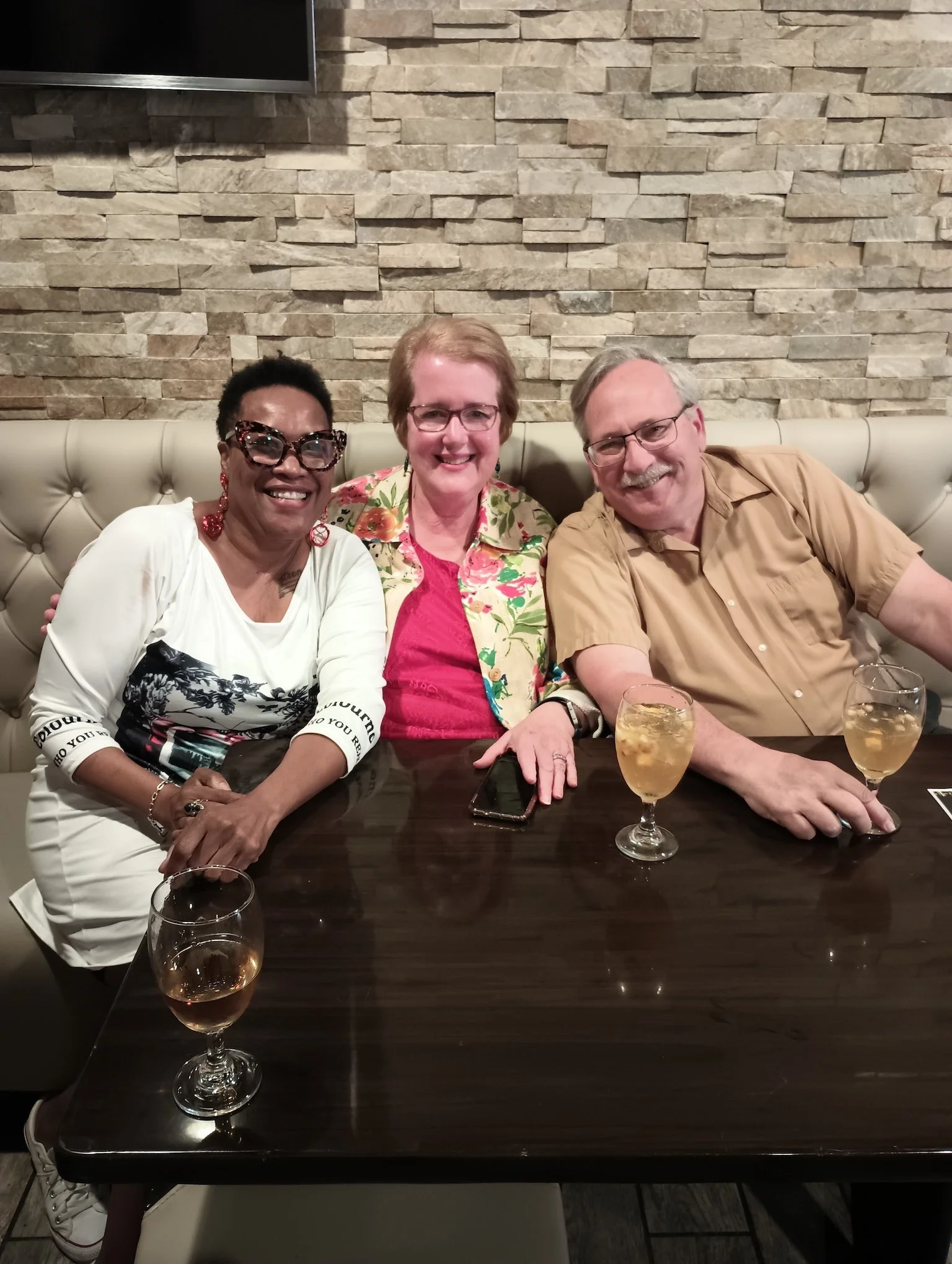 Three people sitting at a dark wooden table in a restaurant, smiling. Two women and one man, with drinks in front of them. The background features a textured stone wall.