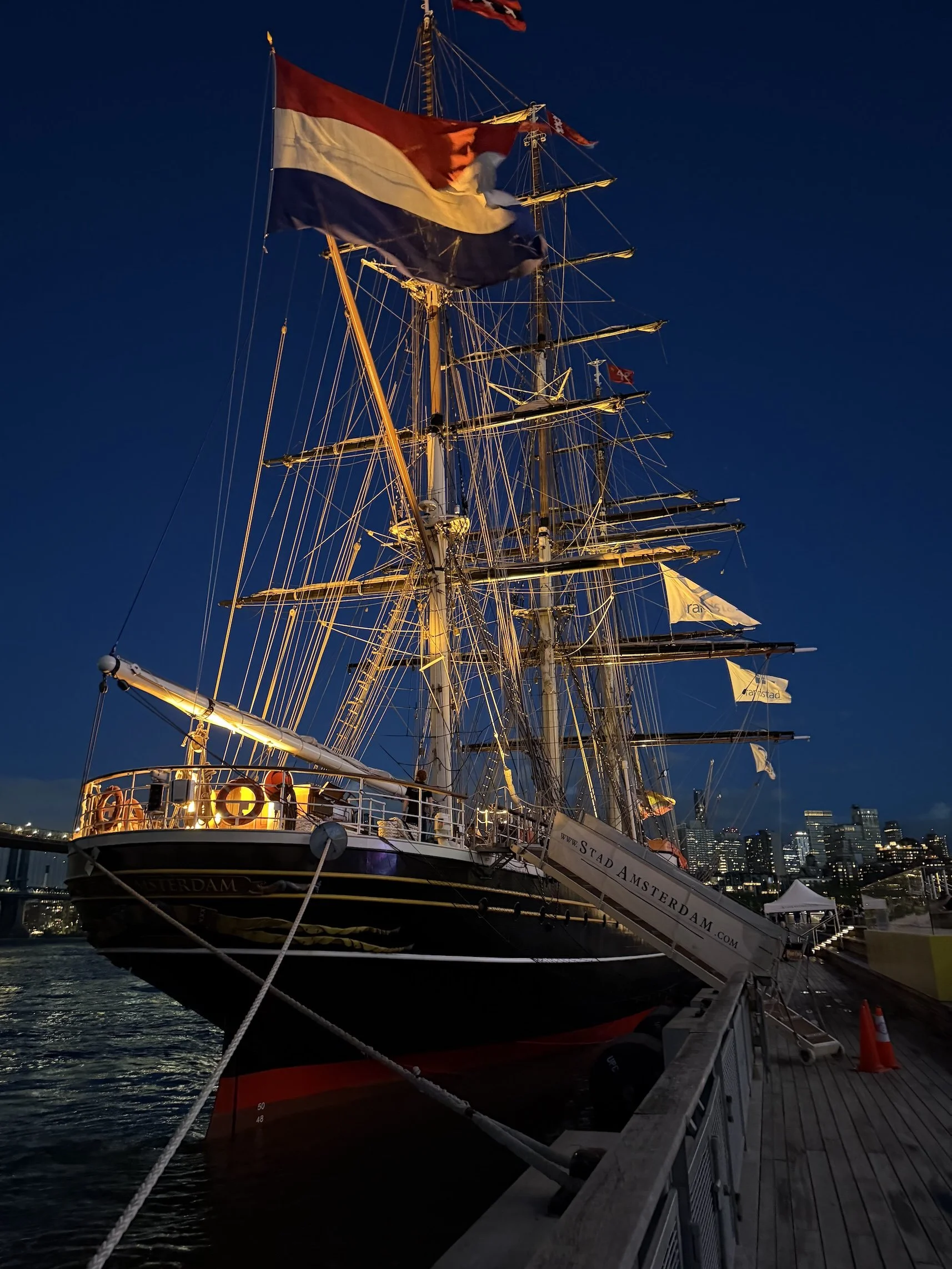 A tall ship with illuminated rigging and flags docked at a harbor during evening with city skyline in the background.