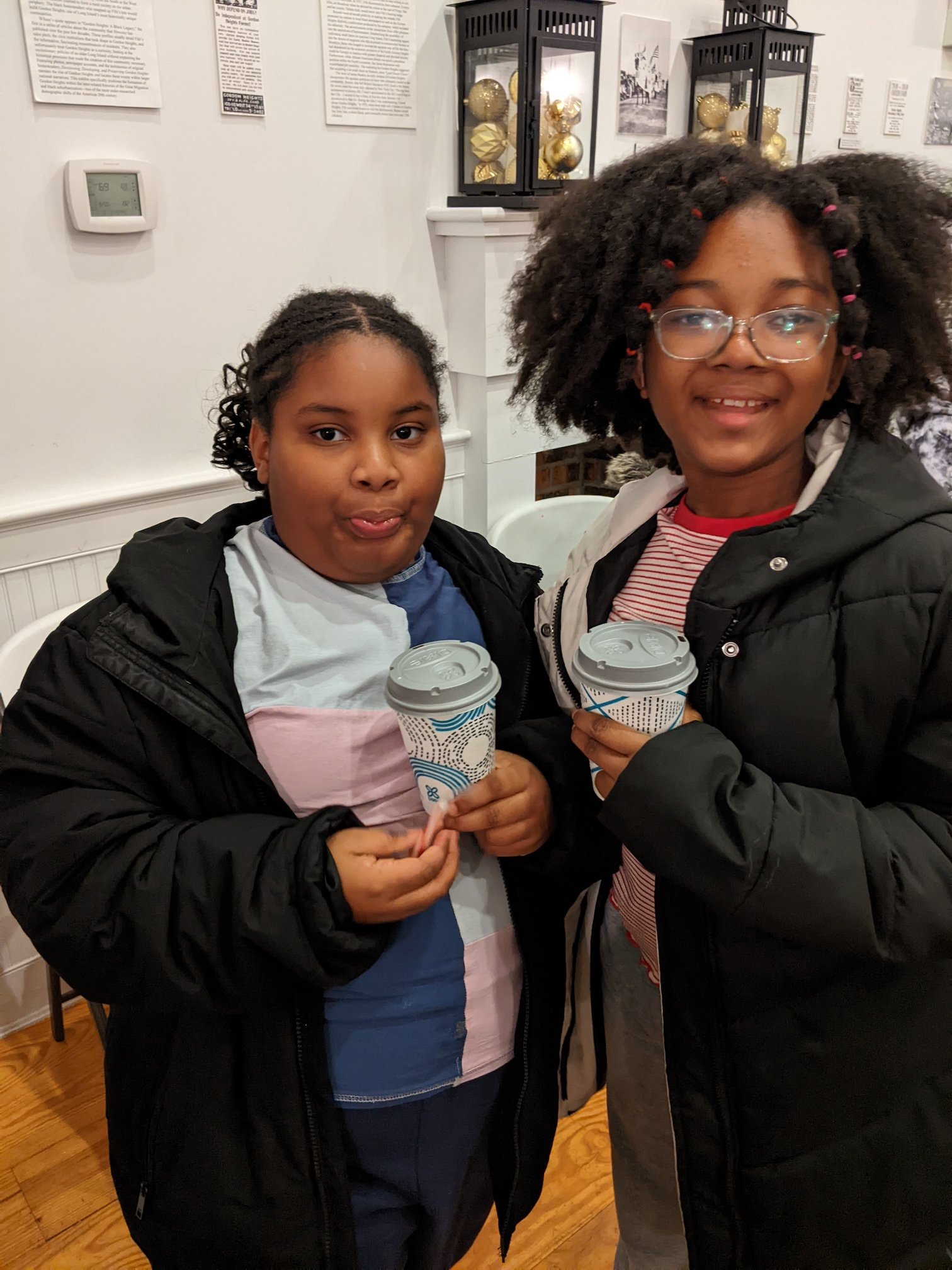 Two young girls holding cups of coffee and smiling inside a room with white walls decorated with framed pictures and Christmas ornaments encased in black lantern-style display cases.