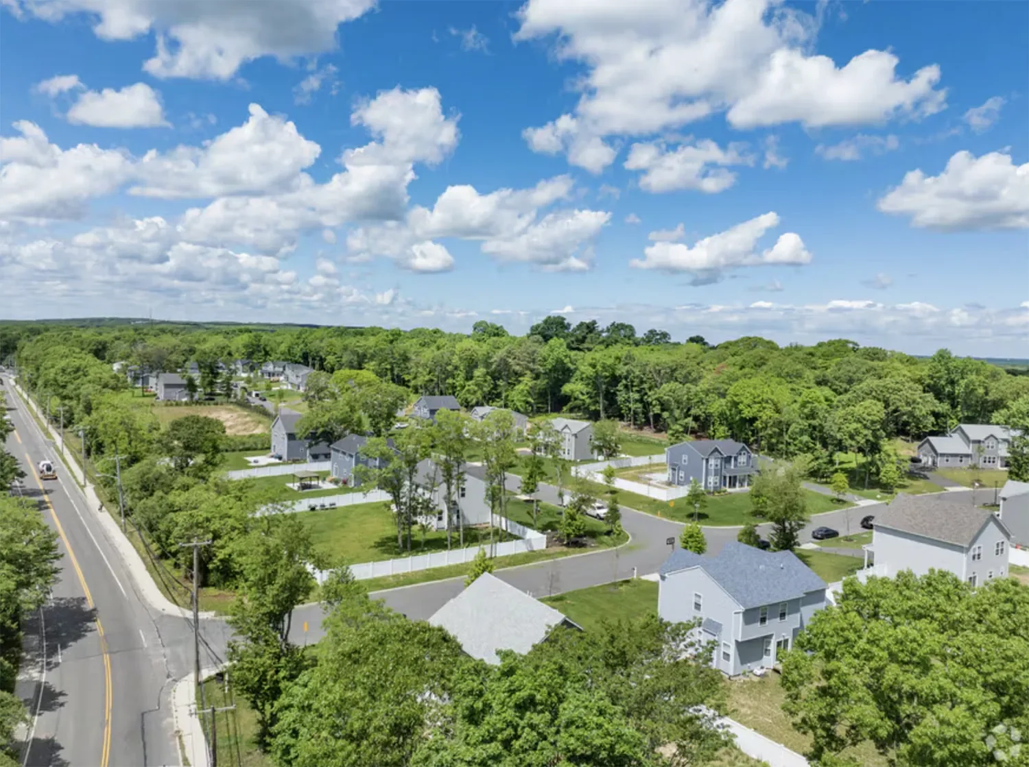 Aerial view of a suburban neighborhood with houses, roads, and lush green trees under a partly cloudy blue sky.