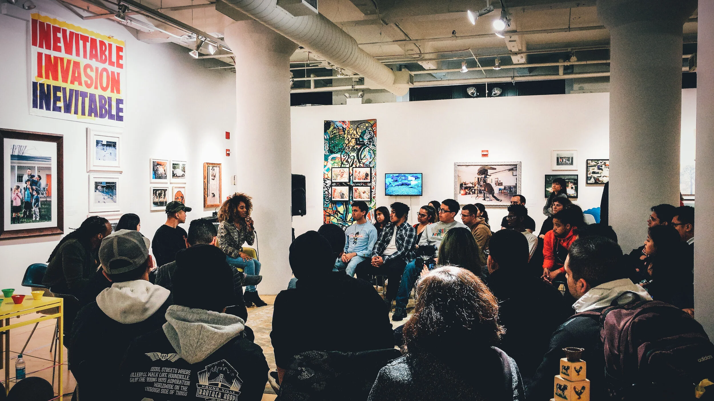 A group of people seated in a gallery space, listening to a woman speaking into a microphone. The gallery has various photos and artwork on the walls, and a large banner reading 'INEVITABLE INVASION' in bold letters.