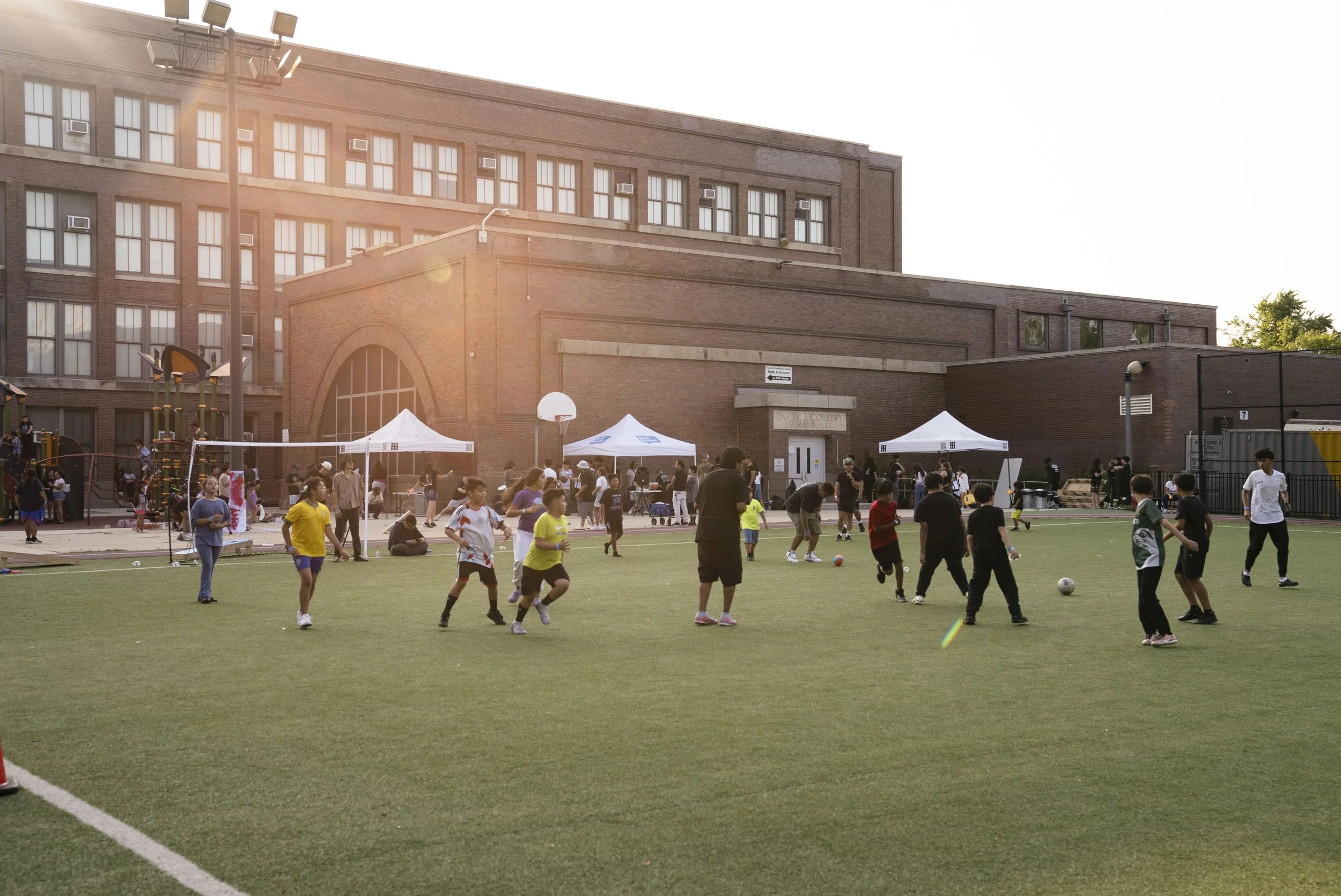 Children and adults playing soccer on a field in front of a school building with tents and playground equipment at sunset.
