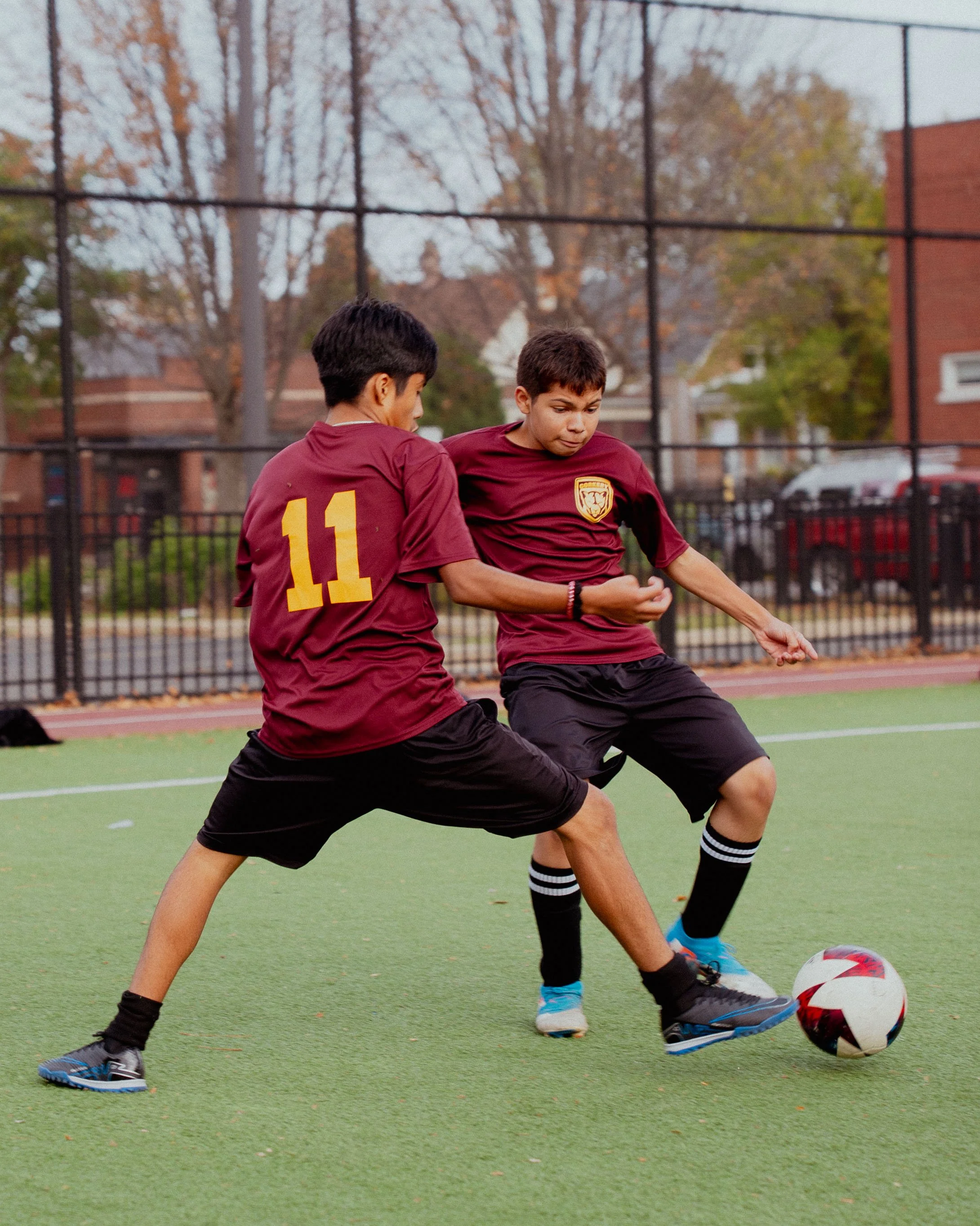 Two boys in maroon soccer jerseys and black shorts playing soccer on an outdoor field, vying for control of the ball. One jersey has the number 11. Trees and a fence are in the background.