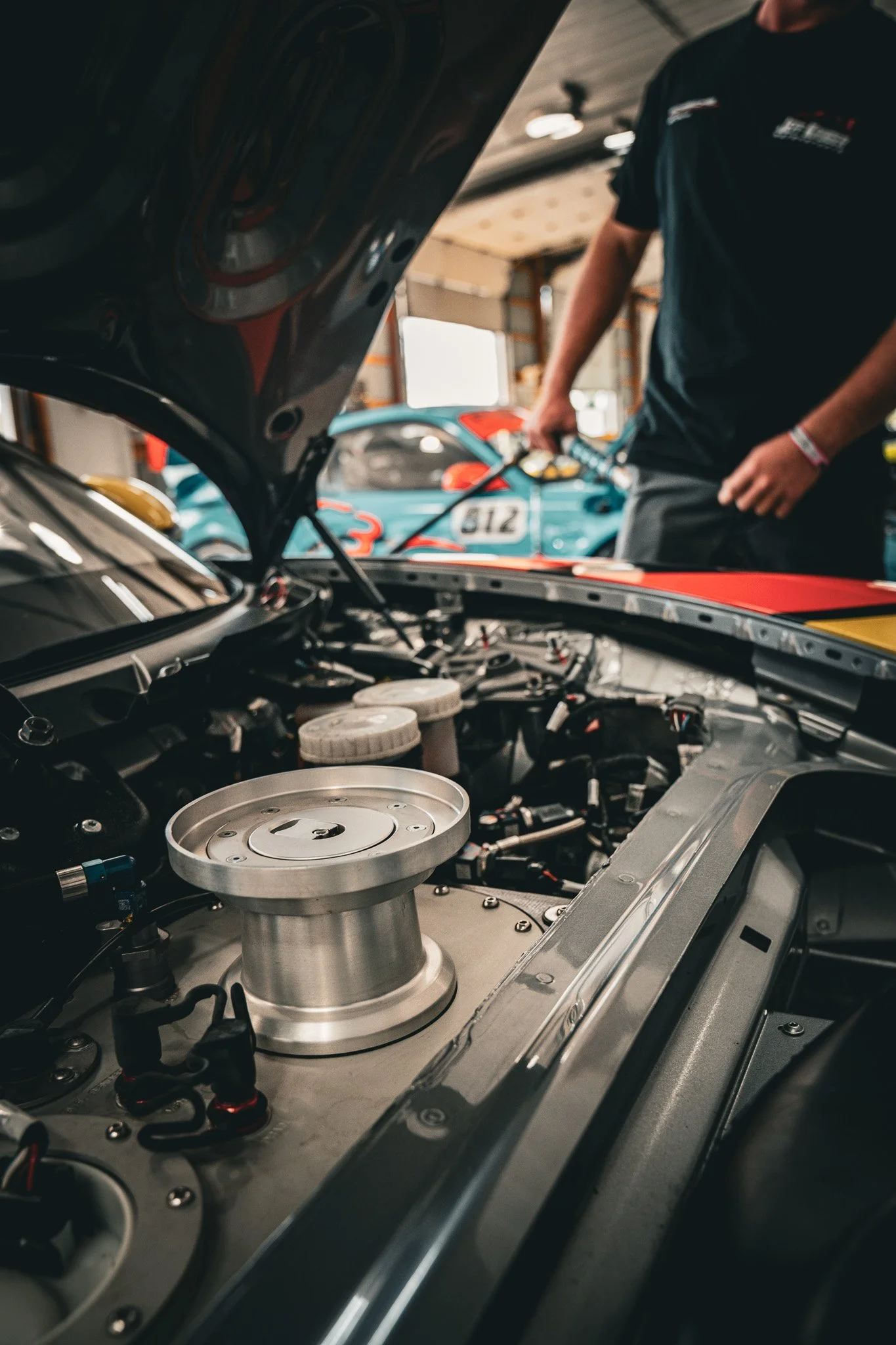 Close-up of a race car engine with a mechanic working in the background at a garage, two race cars with numbers visible in the background.