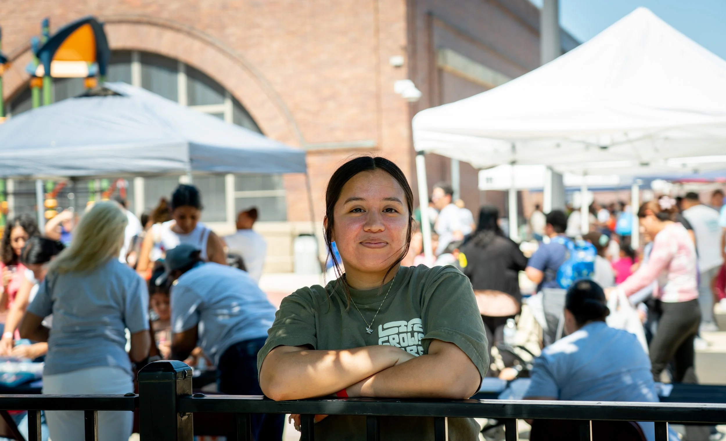 A young woman with long dark hair behind a black railing at an outdoor event, smiling with arms crossed, with tents and a crowd in the background on a sunny day.