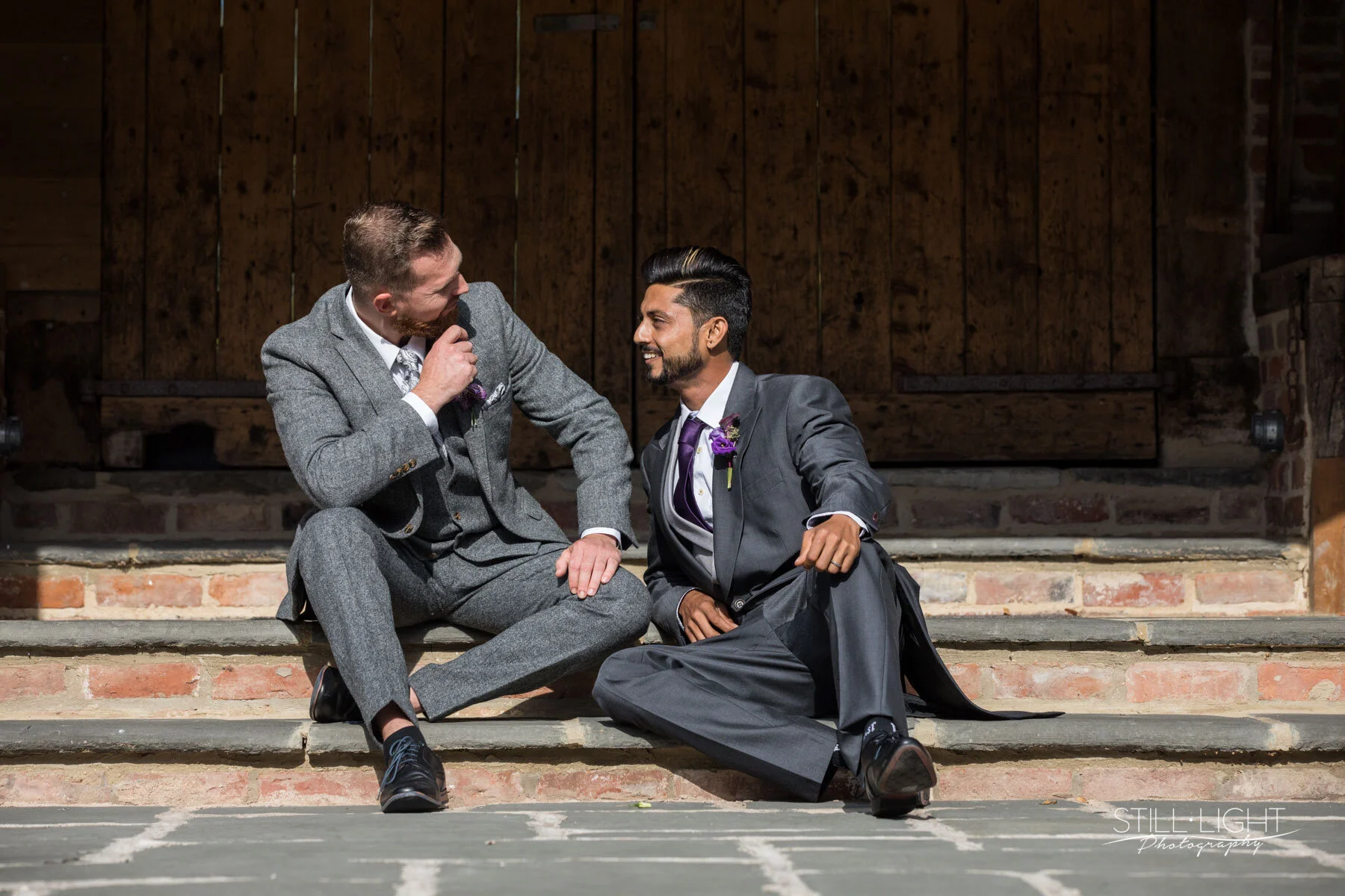 Two groomsmen in grey tailed suits and purple ties and pinholes sat on the steps outside talking