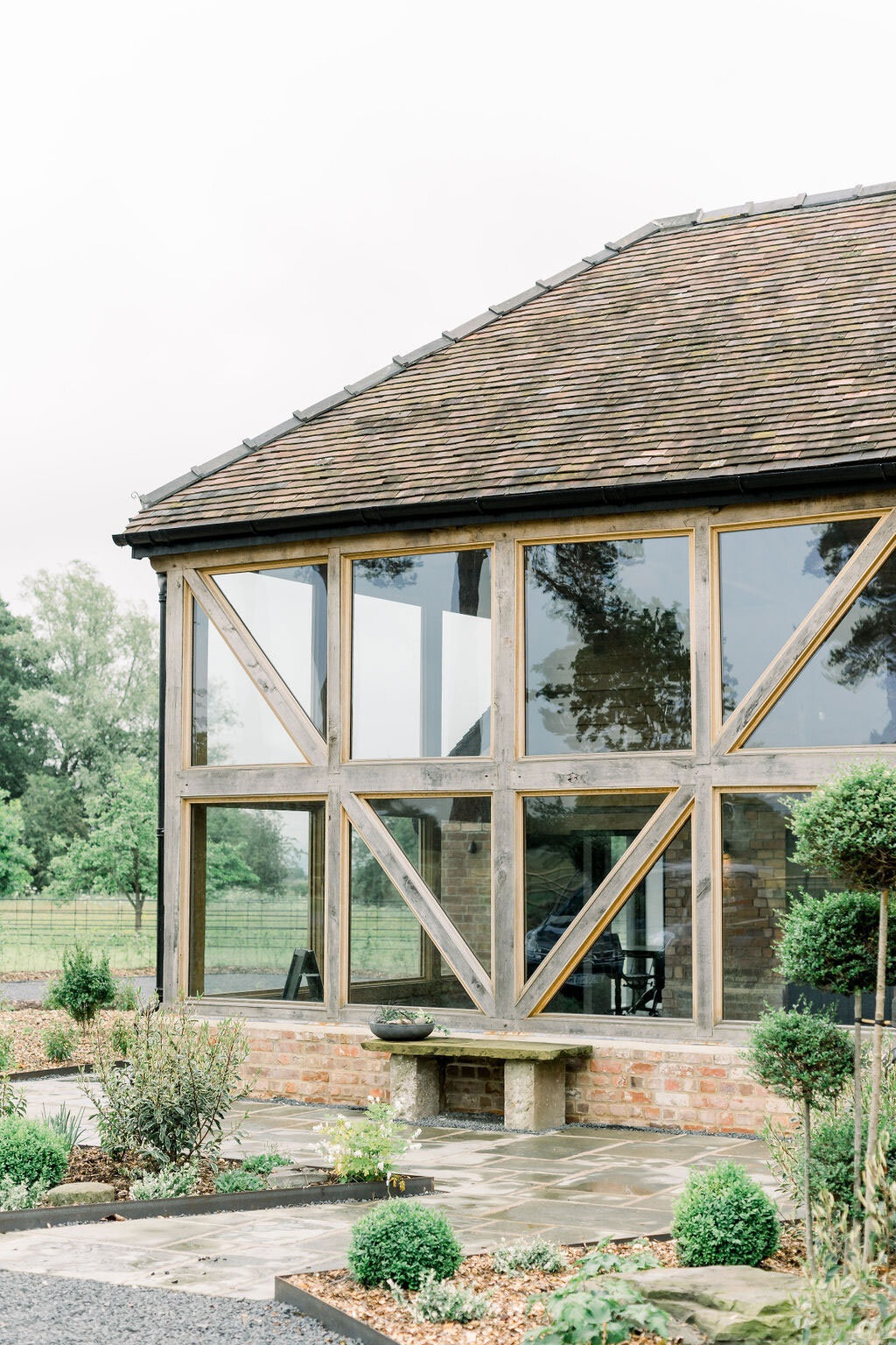 Outside the Cider Barn, with large sqaure panel windows inserted within the beams. Stoneflagged patio walkway and green foliage in boarders