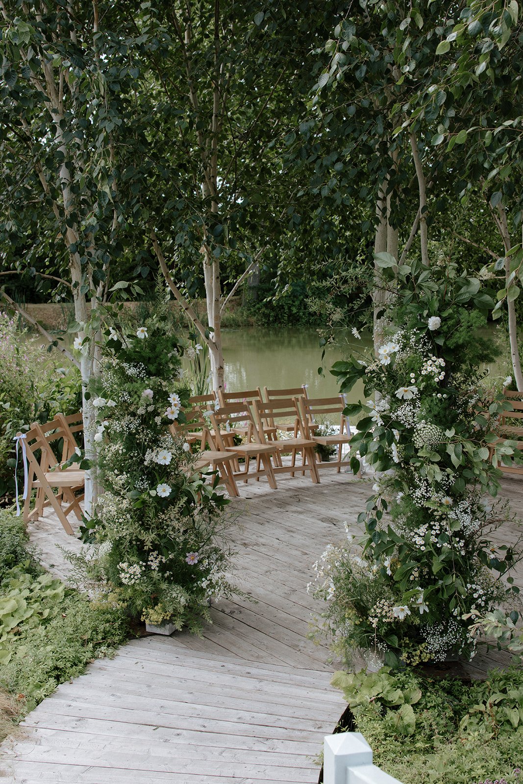 Wedding ceremony on wooden jetty over the lake, wooden chairs arrange beyond a floral archway
