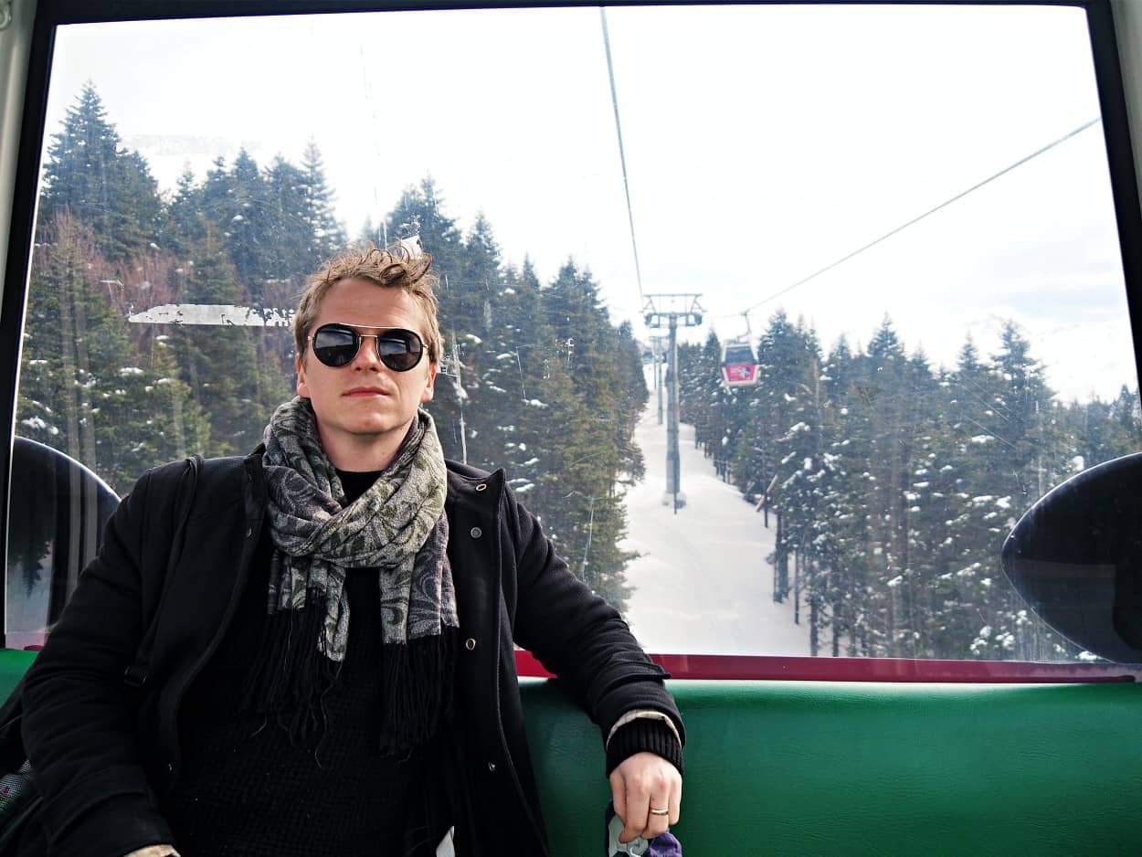 A man wearing sunglasses sitting against the window of  a cable car in Bursa. The forest, the cable car and the snow are in the background