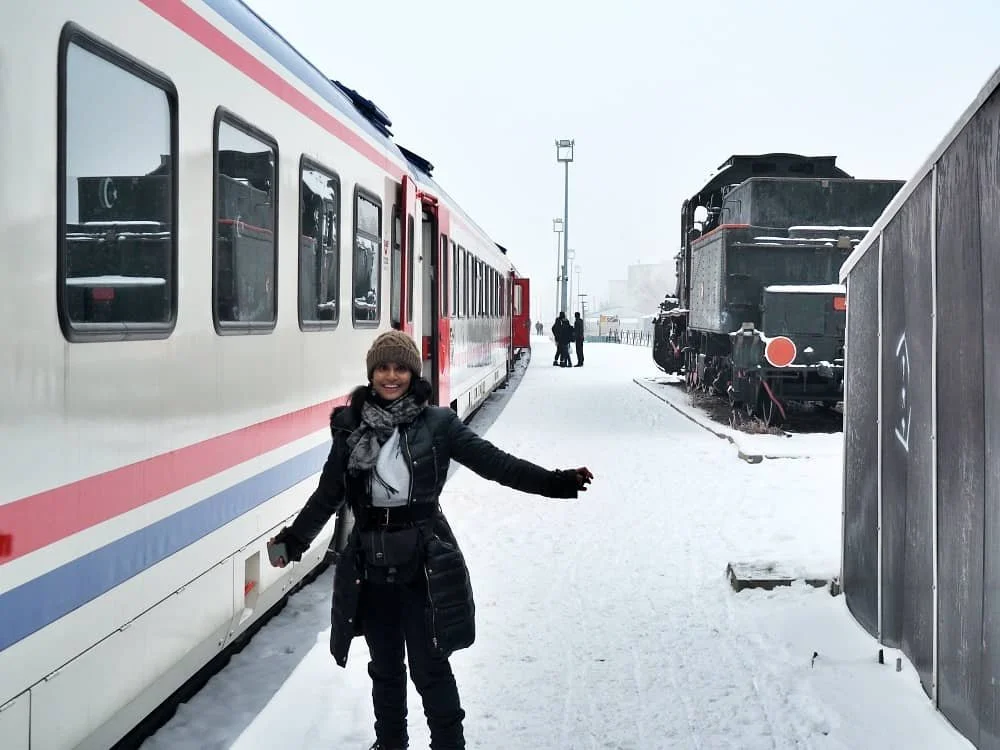 A woman dressed in winter clothing poses for a photograph in front of the Dogu Express train on a snowy platform, ankara kars train