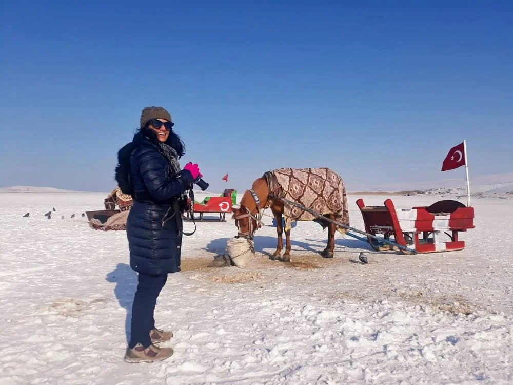 Sasha holding her camera and smiling at the camera in front of a horse and a horse-drawn sleigh on Lake Cildir in Kars, Turkey