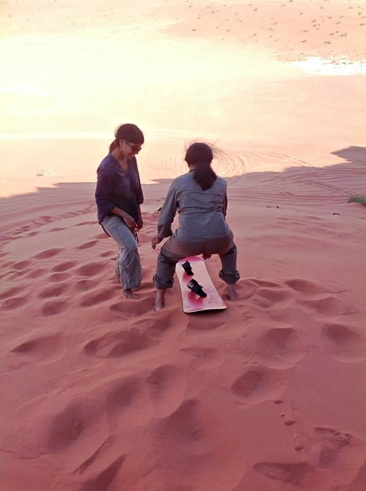 Sasha's mum squats down onto a board with help from Sasha to try out sand boarding in the dessert in Wadi Rum