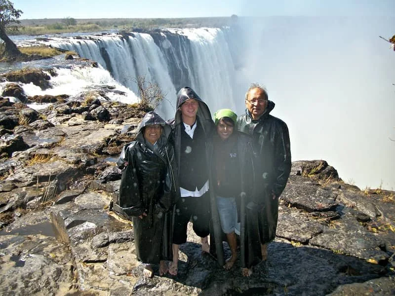 Sasha, Vincent and her mum and dad wear long raincoats at the edge of Victoria Falls on Livingstone Island in Zambia