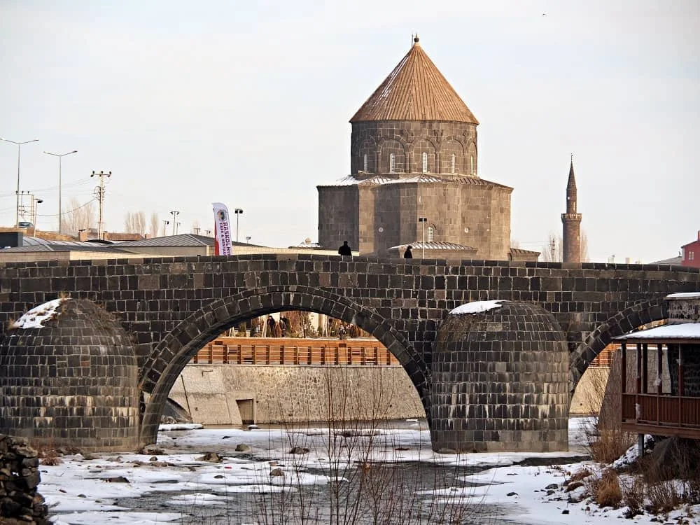 Tas Kopru, Kars' stone bridge over a partially frozen Kars river, with Kumbet mosque in the background.