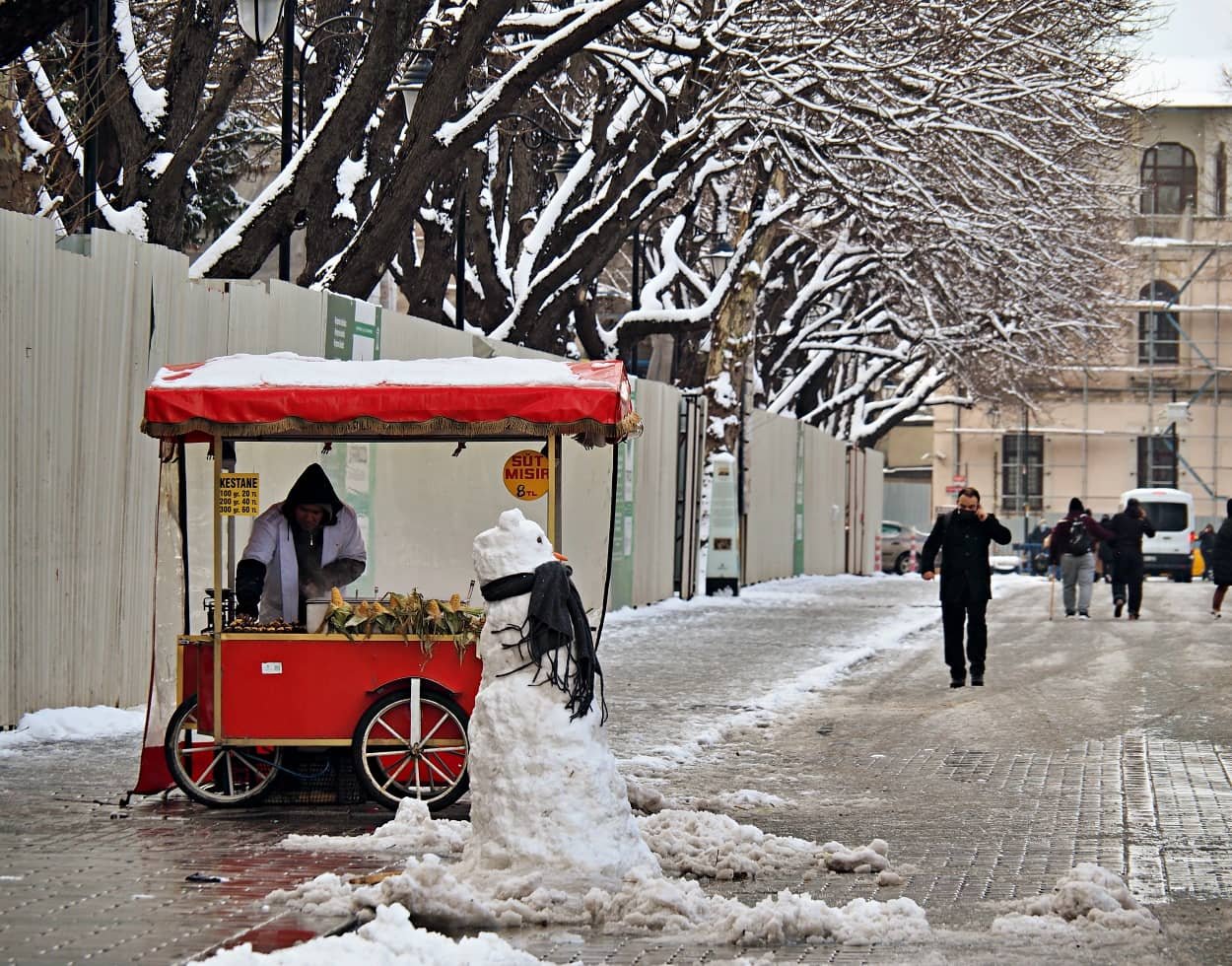 A man selling corn and chestnuts from a bright red cart in the snow iwith a snowman dressed in a car next to him. In the background the trees are covered in snow. Winter in istanbul temperature