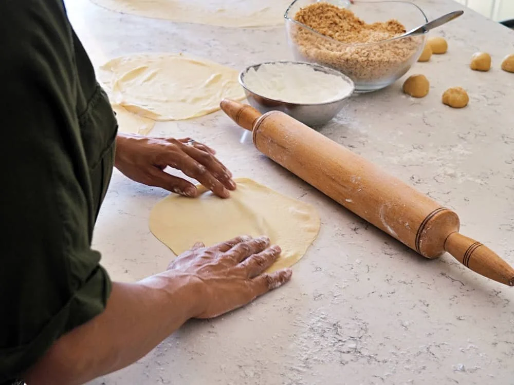 close up of hands rolling out baklava pastry with a rolling pin with crushed walnuts, flour and balls of dough in the background