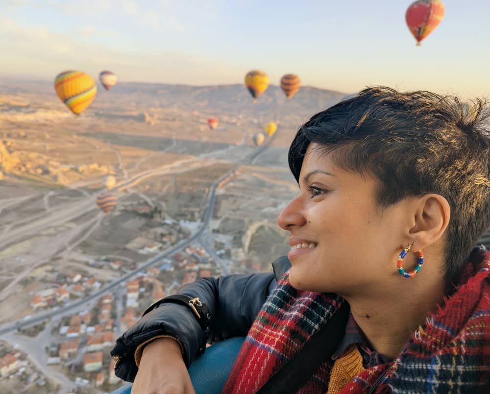 A woman with short black hair looks over the edge of a hot air balloon with other balloons in the background in Cappadocia Turkey.